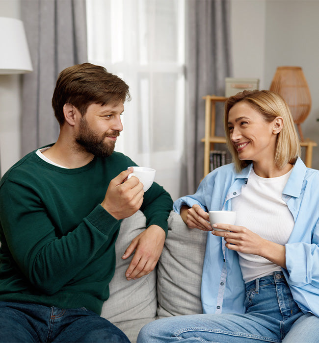 Man and woman sitting on a couch, holding coffee cups and smiling at each other in a cozy living room.